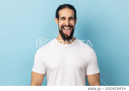 Portrait of comic positive man with beard wearing white T-shirt looking cross-eyed, having fun with silly face expression, showing tongue out. Indoor studio shot isolated on blue background. Portrait of comic positive man with beard wearing white T-shirt looking cross-eyed, having fun with silly face expression, showing tongue out. Indoor studio shot isolated on blue background. 102429434
