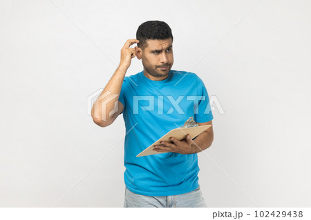 Portrait of pensive puzzled unshaven man wearing blue T- shirt standing with clipboard, writing something on paper, thinking about plan. Indoor studio shot isolated on gray background. Portrait of pensive puzzled unshaven man wearing blue T- shirt standing with clipboard, writing something on paper, thinking about plan. Indoor studio shot isolated on gray background. 102429438