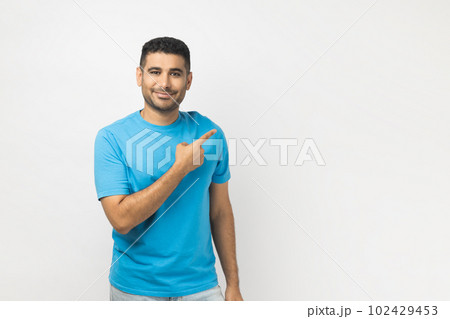 Portrait of smiling cheerful attractive unshaven man wearing blue T- shirt standing pointing copy space for advertisement or promotional text. Indoor studio shot isolated on gray background. Portrait of smiling cheerful attractive unshaven man wearing blue T- shirt standing pointing copy space for advertisement or promotional text. Indoor studio shot isolated on gray background. 102429453