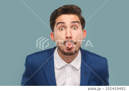 Portrait of funny handsome man with mustache standing sticking tongue out, showing childish behavior, wearing white shirt and jacket. Indoor studio shot isolated on light blue background. 102429482