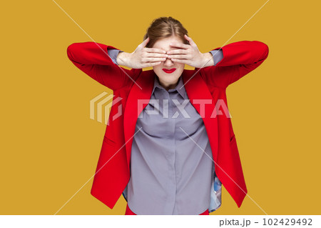 Portrait of shocked woman with red lips standing covering her eyes with palm, do not want to see something scary, wearing red jacket. Indoor studio shot isolated on yellow background. 102429492