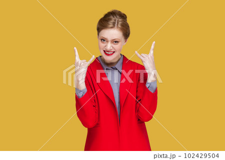 Portrait of excited crazy woman with red lips standing showing rock and roll gesture, having fun on festival, wearing red jacket. Indoor studio shot isolated on yellow background. Portrait of excited crazy woman with red lips standing showing rock and roll gesture, having fun on festival, wearing red jacket. Indoor studio shot isolated on yellow background. 102429504