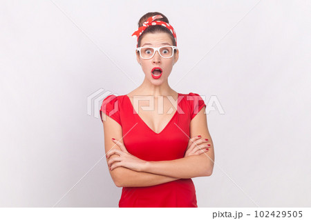 Portrait of shocked surprised woman wearing red dress, glasses and head band standing with crossed arms, looking at camera with big eyes and open mouth. Indoor studio shot isolated on gray background. Portrait of shocked surprised woman wearing red dress, glasses and head band standing with crossed arms, looking at camera with big eyes and open mouth. Indoor studio shot isolated on gray background. 102429505