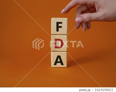 FDA - Food Drug Administration symbol. Wooden cubes with word FDA. Doctor hand. Beautiful orange background. Medical and Food Drug Administration concept. Copy space. FDA - Food Drug Administration symbol. Wooden cubes with word FDA. Doctor hand. Beautiful orange background. Medical and Food Drug Administration concept. Copy space. 102429921