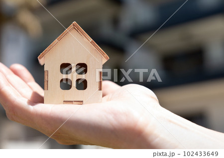 Wooden Model of a country house on a woman's palm with a residential building on the background 102433649