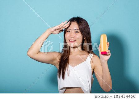 Beautiful Asian young woman holding body lotion on hand in studio shot isolated on blue background, Brunette female hold cream sunblock bottle, tube of sunscreen, Beauty cosmetics facial treatment 102434456