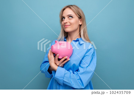dreaming blond young adult hugging a piggy bank where she keeps savings on a blue isolated 102435709