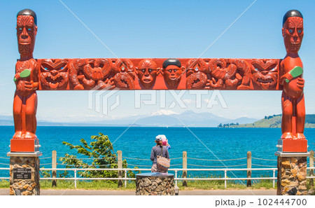Unidentified woman with her baby sitting under the traditional Maori wood carving and looking to Tongariro volcano through lake Taupo. Unidentified woman with her baby sitting under the traditional Maori wood carving and looking to Tongariro volcano through lake Taupo. 102444700