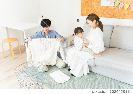 リビングで妻が子供の面倒を見ている間、洗濯物をたたんで家事に協力している夫 102445038