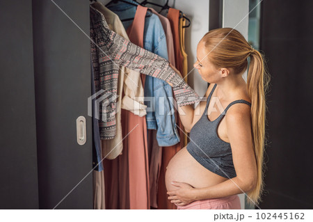 A pregnant woman has nothing to wear. A pregnant woman stands in front of a closet with clothes and does not know what to wear because the clothes do not fit on her 102445162