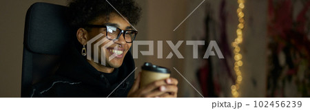 Leisure activities. Happy young guy with piercing wearing glasses holding disposable cup of coffee, sitting at the table and watching videos using laptop late at night 102456239