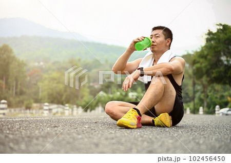 Sweaty and thirsty mature Asian man sits on the road in a public park and drinks water Sweaty and thirsty mature Asian man sits on the road in a public park and drinks water 102456450