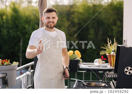 Portrait of handsome young chief in apron stand by the BBQ grill and smile. Male look into camera and showing thumbs up. 102458292