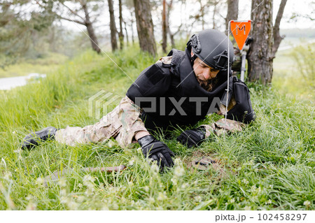 A man in a military uniform and a bulletproof vest works in the forest to demine the territory. A man warns of danger by making a red mark 102458297
