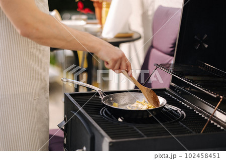 Middle selection of human hands stirs with a wooden spoon food on frying pan. BBQ grill outdoor on fresh air 102458451