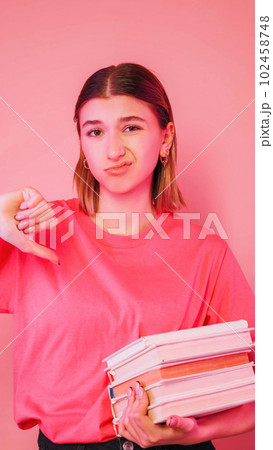 Dislike student. Boring education. Exam failure. Dissatisfied sad girl holding stack of books showing thumb down sign grimacing isolated on neon color pink background. 102458748