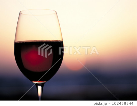 赤ワインの入ったワイングラスと夕景のアンバーな空に浮かぶ太陽のシルエット 102459344