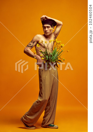 Full-length portrait of stylish, handsome, young man posing shirtless in beret and beige pants against yellow studio background 102460504