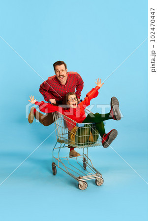 Portrait with couple, man and pretty woman having a shopping cart race over blue color studio background. Adventure time 102470447