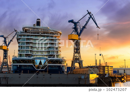 Scenic back view large modern luxury cruise ship liner under construction building cranes at dry dock shipyard in Hamburg port dramatic sunset sky background. Big vessel manufactoring site industry 102473207