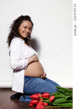 African American curly haired happy adult pregnant woman, holding hands on her belly, smiling looking at camera, sitting on wooden floor, isolated over white background. 9 month. Pregnancy. Maternity 102478714