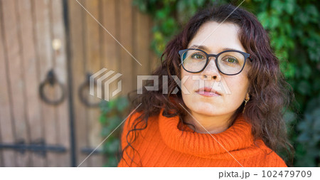 Banner Autumn portrait of an attractive young woman in stylish glasses in a knitted fashionable orange sweater on background of leaves and big door copy space . Girl walks in city 102479709