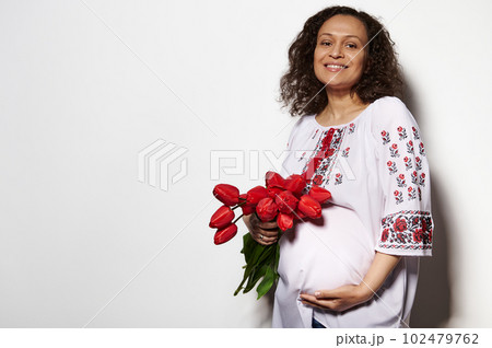 Delightful adult pregnant woman, expectant mother dressed in Ukrainian ethnic embroidered clothing, holding her belly, posing with a bouquet of red tulips, isolated on white background. Copy ad space 102479762
