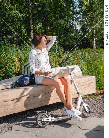 Woman sits with laptop on urban park bench. Freelancer at work. Student learns remotely from outdoors. Modern lifestyle. Summer vibes. Outdoor workplace. Woman sits with laptop on urban park bench. Freelancer at work. Student learns remotely from outdoors. Modern lifestyle. Summer vibes. Outdoor workplace. 102484954