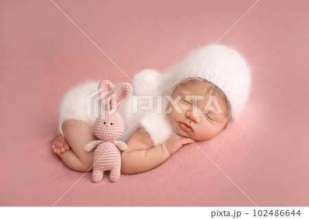 Top view of a newborn baby girl sleeping in a white overalls, with a white cap on her head, on a pink background. With pink toy rabbit. Beautiful portrait of a small newborn baby 7 days, one week old. Top view of a newborn baby girl sleeping in a white overalls, with a white cap on her head, on a pink background. With pink toy rabbit. Beautiful portrait of a small newborn baby 7 days, one week old. 102486644