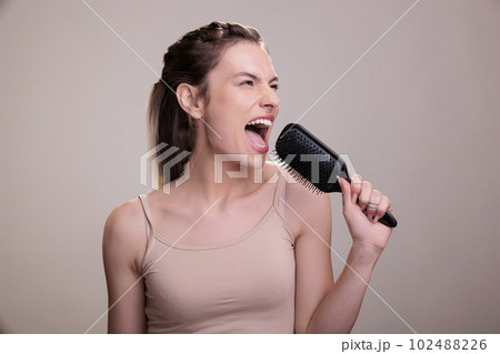 Beautiful woman singing loudly in hairbrush during hairstyle routine. Attractive young caucasian model posing with comb, using hairdressing tool as microphone and having fun 102488226