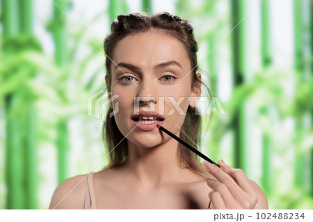 Young model doing lips make up with brush portrait. Attractive woman with healthy fresh skin applying lipbalm, showing makeup daily routine and looking at camera on bamboo background 102488234