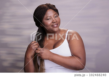 Natural beautiful african american woman making braids hairstyle, wearing white top, smiling and looking at camera. Attractive body positive model showing haircare daily procedure Natural beautiful african american woman making braids hairstyle, wearing white top, smiling and looking at camera. Attractive body positive model showing haircare daily procedure 102488240