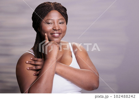 Natural beautiful skincare body positive model touching cheek portrait. African american curvy woman pointing on perfect healthy skin, holding finger on face and looking at camera Natural beautiful skincare body positive model touching cheek portrait. African american curvy woman pointing on perfect healthy skin, holding finger on face and looking at camera 102488242