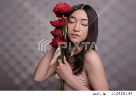 Asian woman with closed eyes holding red poppies branch. Beautiful young model wearing beige casual top, standing with summer flowers bouquet, showing skincare and body care concept 102488244