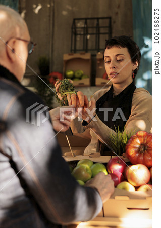 Young woman farmer standing behind farmers market stall showing organic locally grown carrots to customer while selling fresh fruits and vegetables. Mature man buying healthy local food 102488275