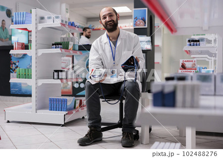 Smiling pharmacist holding medical tonometer ready to measure customers hypertension during checkup visit in pharmacy. Drugstore filled with supplements and drugs, medicine support service 102488276