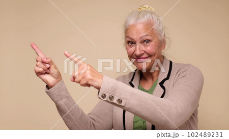 An elderly woman points her finger to attract attention on a beige background An elderly woman points her finger to attract attention on a beige background 102489231