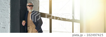 Full length shot of stylish middle aged man in business casual wear and sunglasses leaning on the wall, holding disposable coffee cup while posing in loft interior 102497425