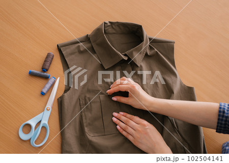 Close up view from above on the hands of a dressmaker, a young woman is manually working on the design of a new collection of khaki shirts. There are threads and scissors on the table nearby 102504141