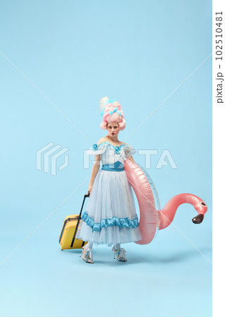 Portrait with young princess, queen wearing dress holding suitcase and inflatable flamingo going to travel over blue background Portrait with young princess, queen wearing dress holding suitcase and inflatable flamingo going to travel over blue background 102510481