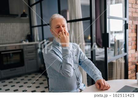 Hungry bearded mature elderly male eating hamburger with beef from fast food restaurant sitting at table and browsing online with laptop computer. Handsome bearded senior man eating delicious burger. 102511425