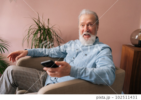 Portrait of happy excited gray-haired mature male holding smartphone using mobile online app, shocked looking at camera with open mouth sitting on armchair. Bearded older man enjoying social media. Portrait of happy excited gray-haired mature male holding smartphone using mobile online app, shocked looking at camera with open mouth sitting on armchair. Bearded older man enjoying social media. 102511431