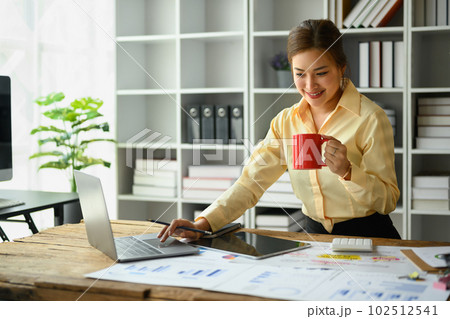 Cheerful asian woman employee drinking coffee and working with statistic reports at office desk 102512541