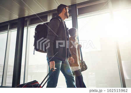The weather is looking great outside. a young couple walking inside of an airport with their luggage and holding hands while looking outside. The weather is looking great outside. a young couple walking inside of an airport with their luggage and holding hands while looking outside. 102515962