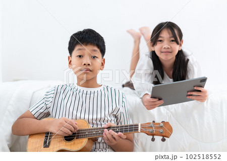 Brother holding an ukulele, sister holding a tablet looking at the camera in the bedroom 102518552