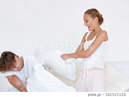 Happy, fun and couple in a pillow fight on the bed in the hotel room on a romantic weekend trip. Happiness, laughing and young woman and man playing together in the morning in the bedroom at home. 102521128