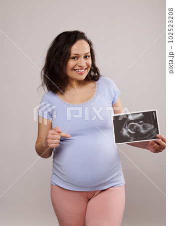 Happy adult pregnant woman, smiling looking at camera, showing her newborn baby sonography and pointing at her belly over isolated isolated white background. Pregnancy. Ultrasound scan image of a baby 102523208