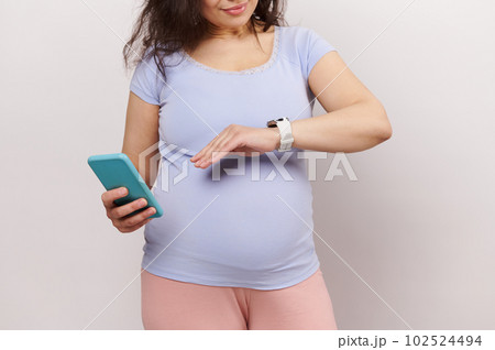 Closeup beautiful pregnant woman holding mobile phone, checking her smart wrist watch, counting baby kicks or movements, making notes in new pregnancy mobile application, isolated on white background 102524494