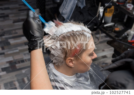The hands of the hairdresser in gloves with a brush paint the hair of the client's head with white paint close-up The hands of the hairdresser in gloves with a brush paint the hair of the client's head with white paint close-up 102527741
