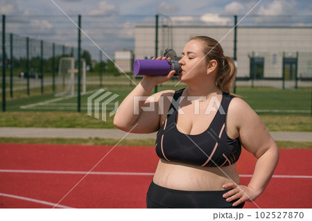 A fat, obese pregnant woman with stretch marks on her stomach drinks water from thirst after fitness classes in the fresh air 102527870